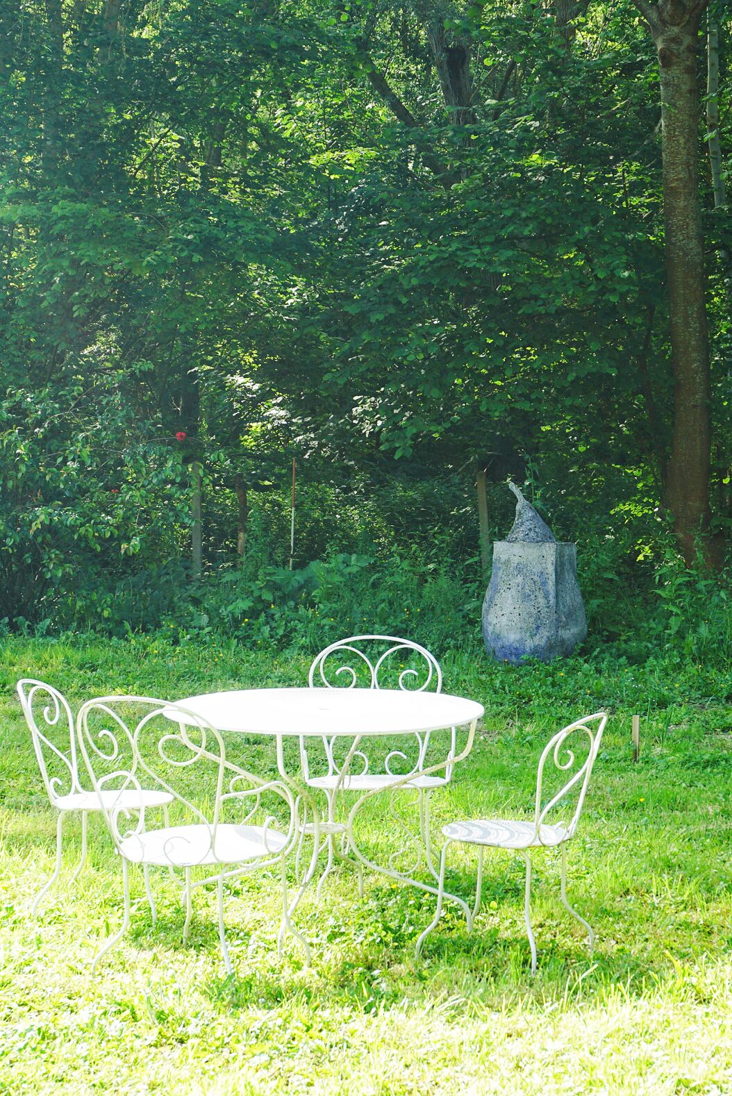 Een witte metalen tafel met stoelen in een grasrijke omgeving, met een grote decoratieve steensculptuur op de achtergrond.