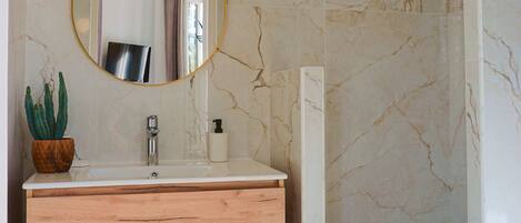 A bathroom with a marble wall, a wooden vanity, a round mirror, a shower, and a potted plant.