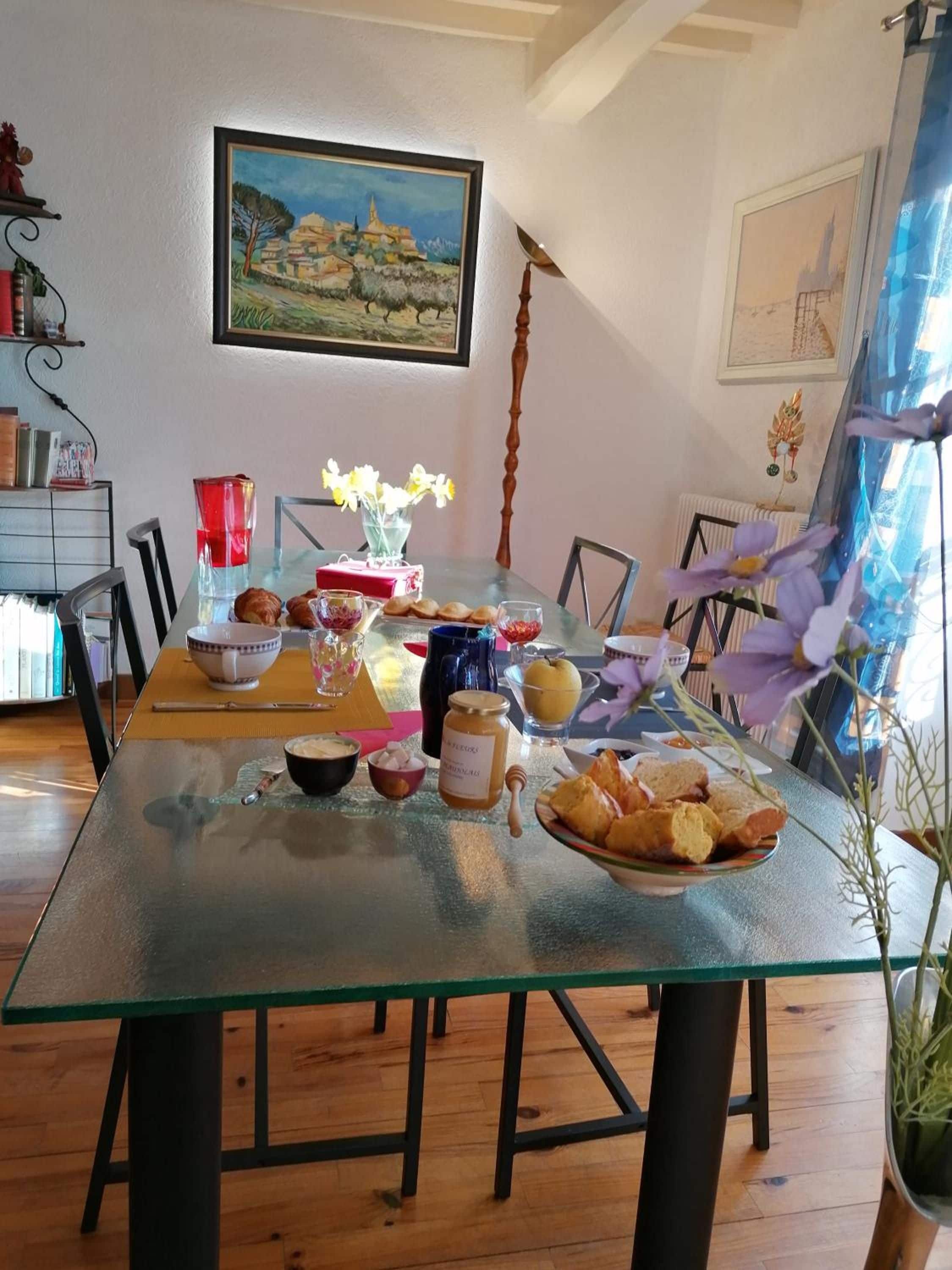 A dining area with a glass table, chairs, a vase with flowers, and a painting on the wall.