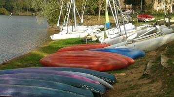 Una hilera de kayaks coloridos alineados en una zona verde a orillas de un lago, con árboles y un cuerpo de agua tranquilo al fondo.