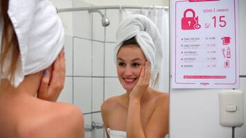 Una persona con una toalla en la cabeza, de pie frente a un espejo en un baño. Un cartel en la pared enumera artículos a la venta y alquiler, incluyendo cerraduras, toallas, cepillos de dientes, pasta de dientes y champú.