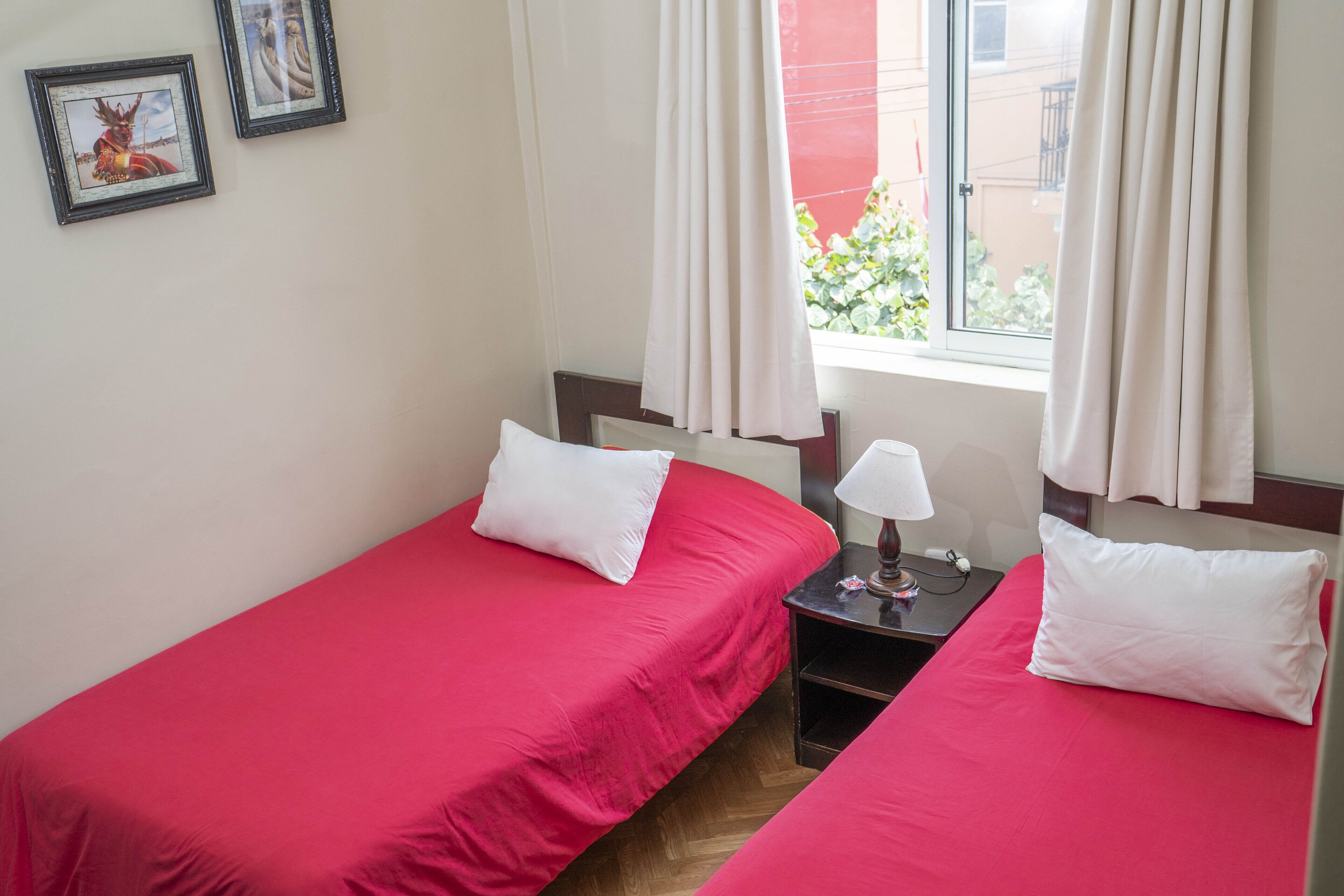 A hotel room with two single beds, each with a white pillow and a red bedspread. There is a nightstand with a lamp and a small table between the beds. The room has a window with white curtains, and two framed pictures on the wall.