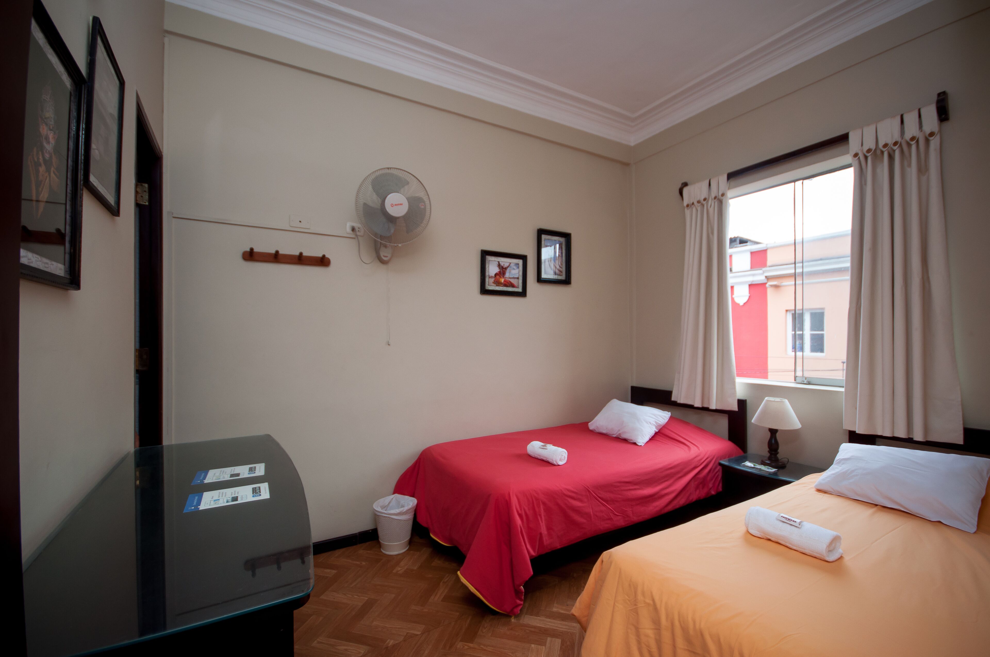 Ein Hotelzimmer mit zwei Betten, einem Schreibtisch, einem Ventilator und einem Fenster mit Vorhängen.