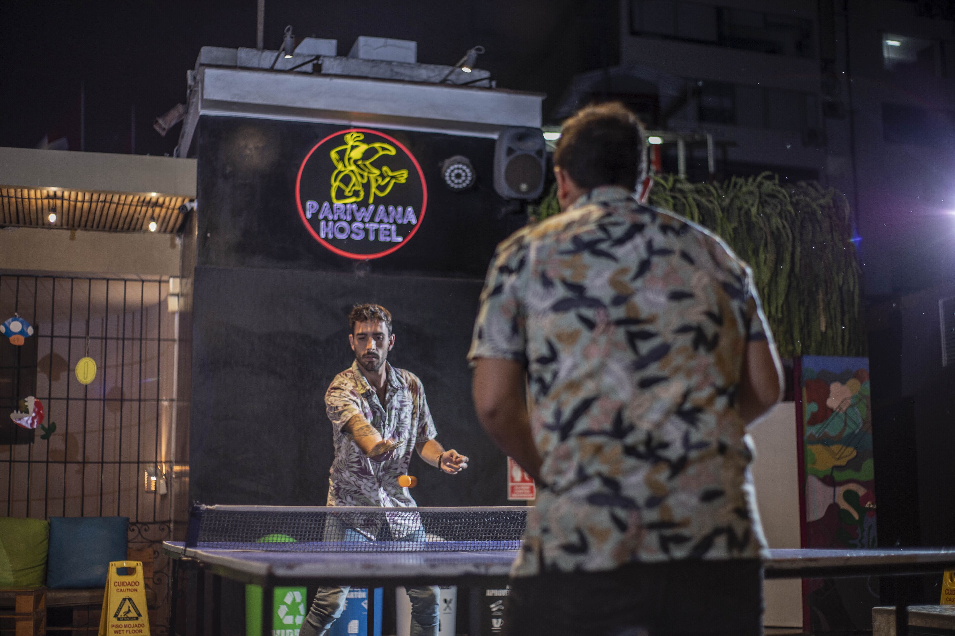 A table tennis match in front of a hostel with a sign reading "PARIWANA HOSTEL".