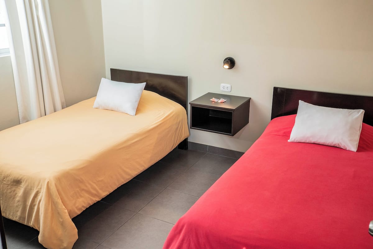 A hotel room with two beds, one with a yellow bedspread and the other with a red bedspread, a small bedside table with a lamp, and a wall-mounted light fixture.