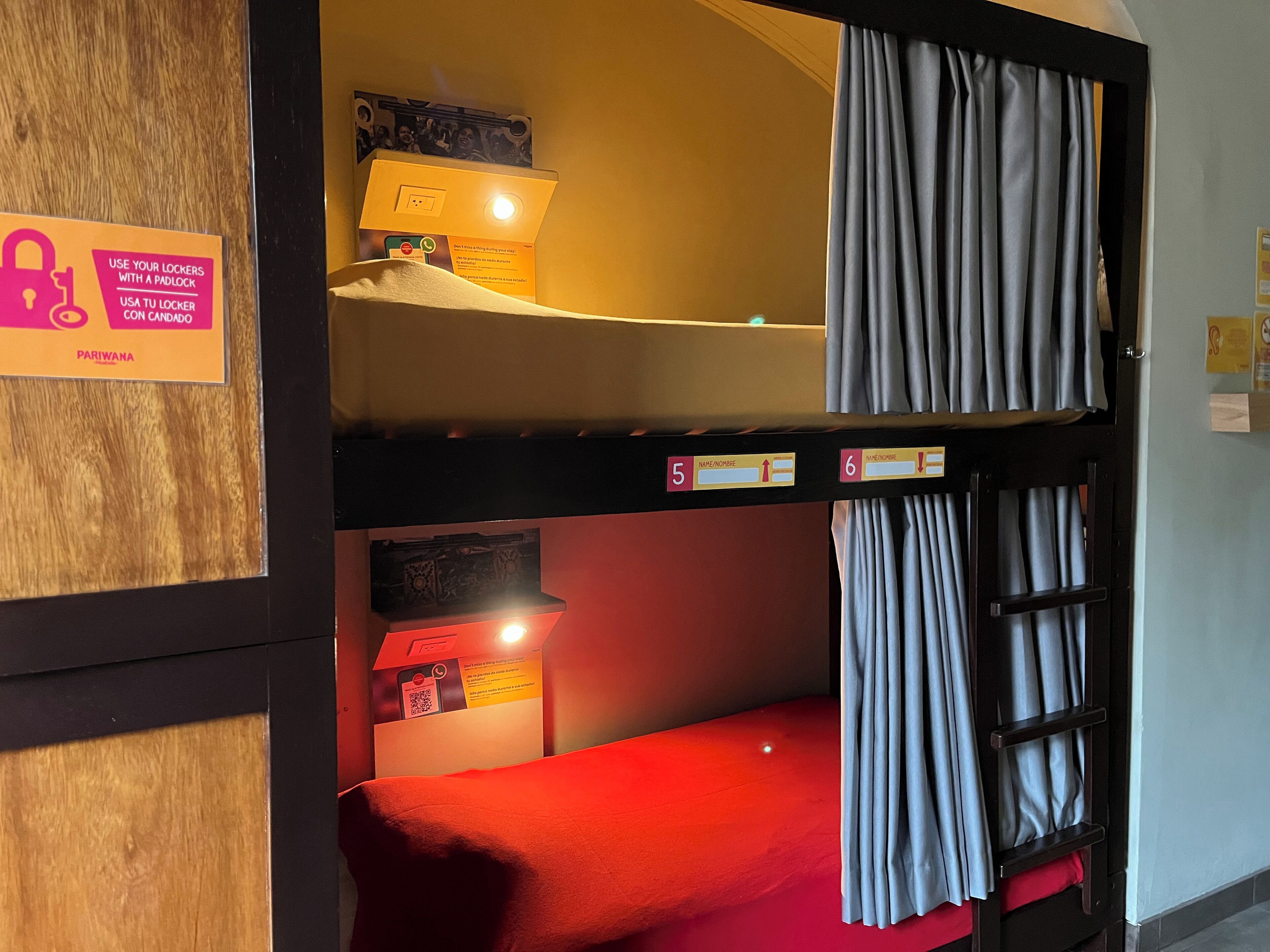 A bunk bed room with a sign instructing to use lockers with a padlock, a lamp on the upper bunk, and a red bedspread.
