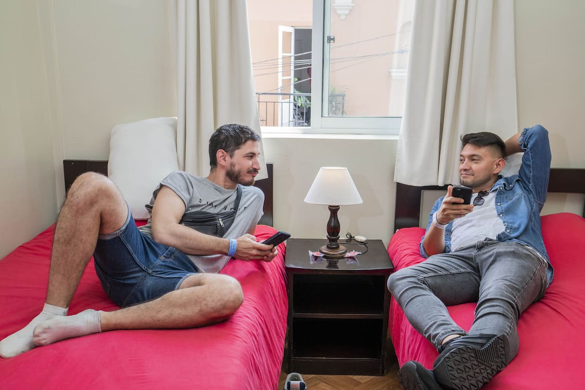 Two individuals sitting on a red bed in a room with a window, a lamp, and a small table.