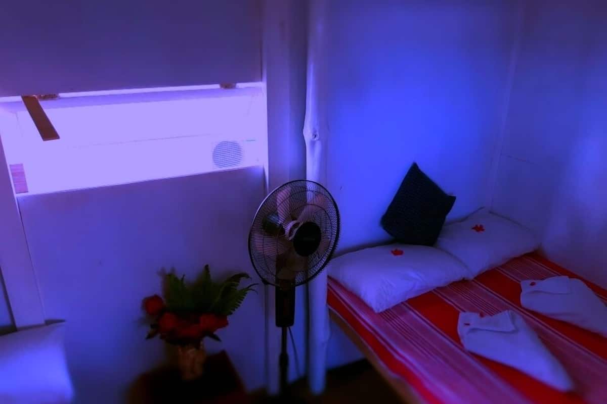 A small, dimly lit room with a single bed, a bedside table with a fan, and a window with a light fixture.