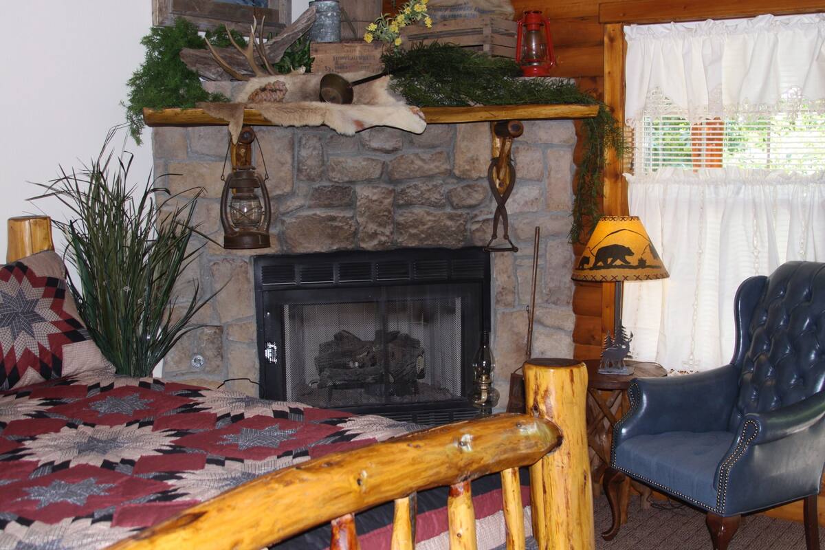 A cozy room with a stone fireplace, a bed with a patterned blanket, a blue leather armchair, and a wooden shelf with decorative items.