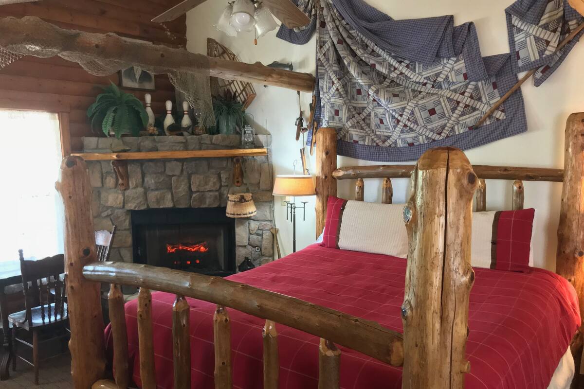 A rustic bedroom with a stone fireplace, wooden bed frame, and a red bedspread.