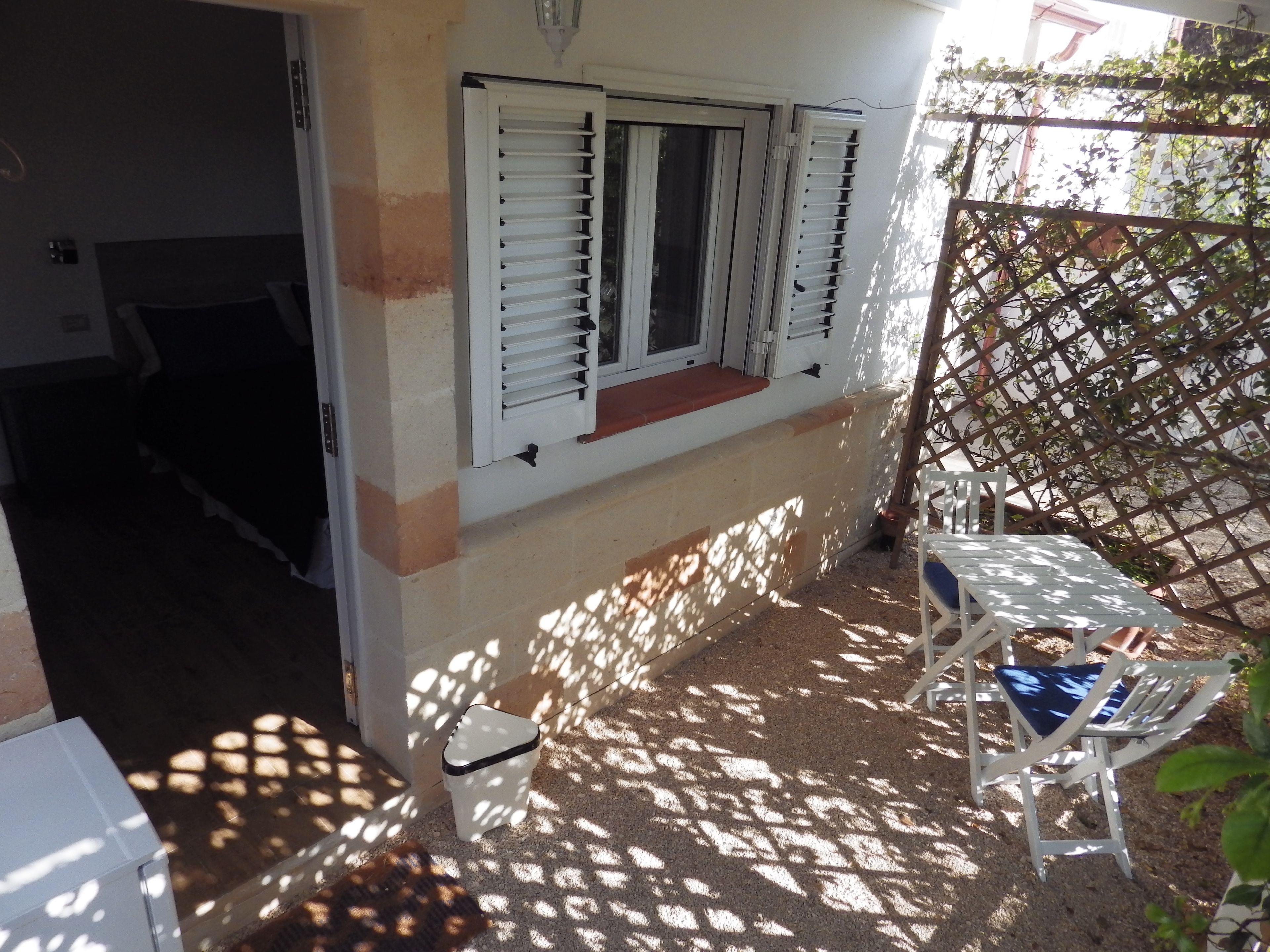 A small outdoor patio area with a white table and chairs, a white trash bin, and a window with white shutters.