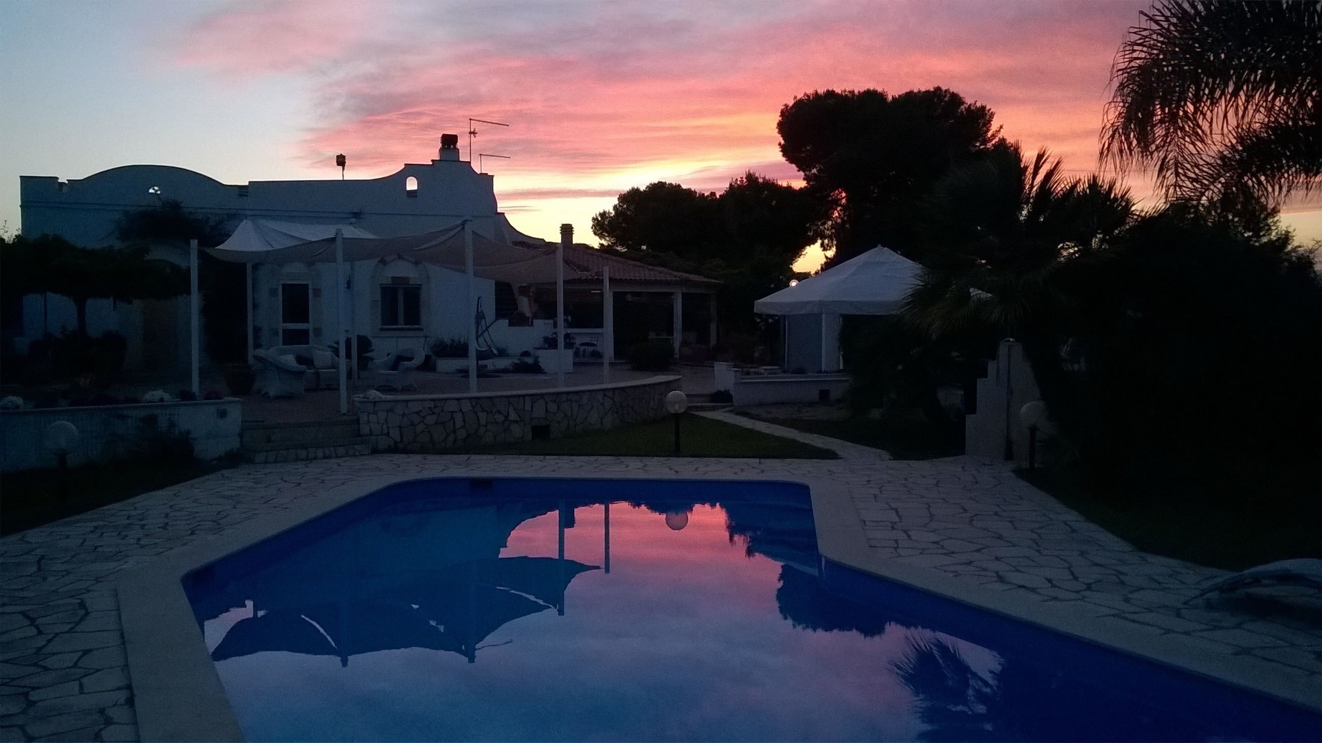 Ein Pool mit spiegelklarer Wasseroberfläche, umgeben von einer Steinterrasse und einem weißen Gebäude mit überdachter Terrasse.