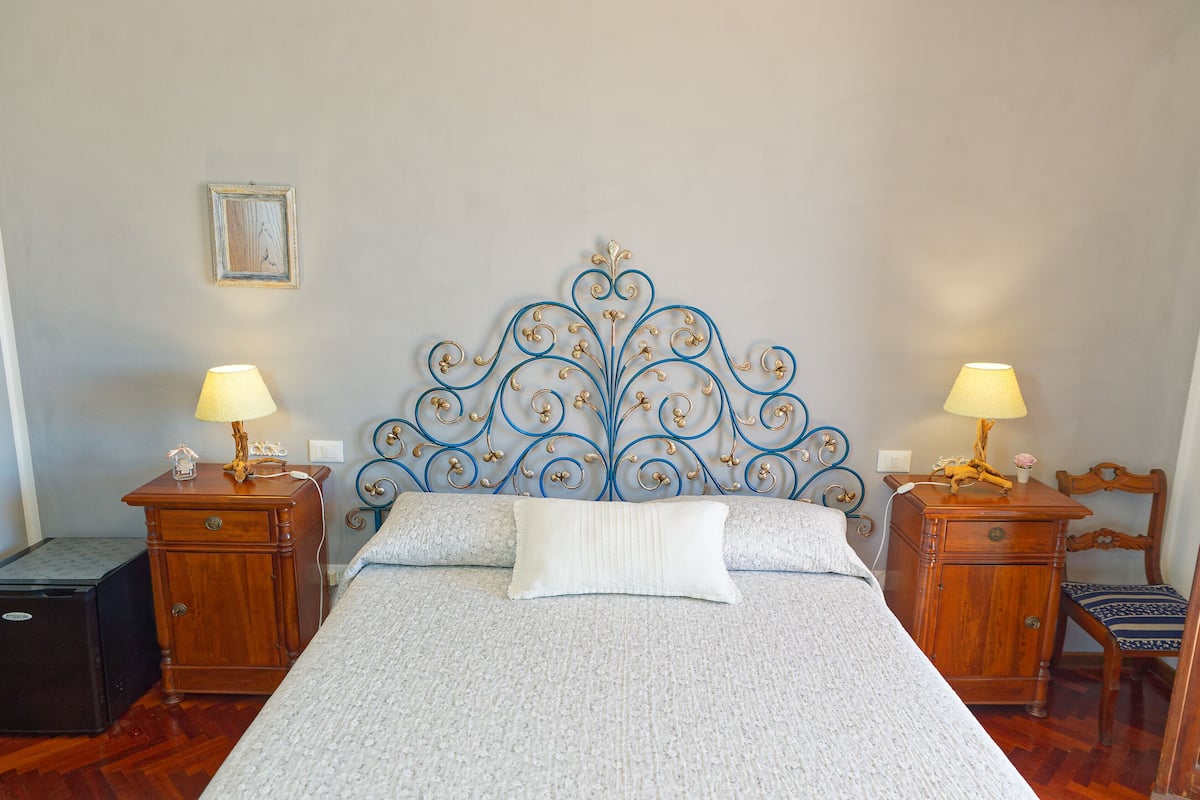 Un lit bien fait, agrémenté d’une tête de lit décorative en métal, deux tables de chevet avec des lampes, une petite armoire en bois et un tableau encadré accroché au mur.