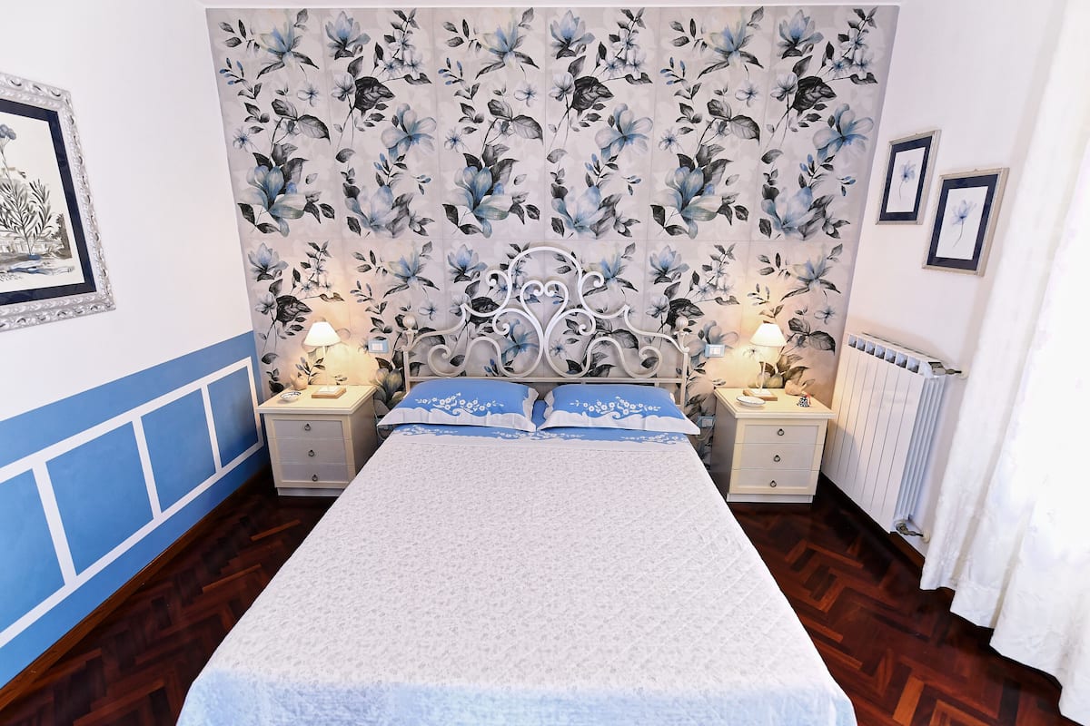 Une chambre à coucher avec une tête de lit à motifs floraux, un lit avec une literie blanche, deux tables de chevet avec des lampes et un radiateur.