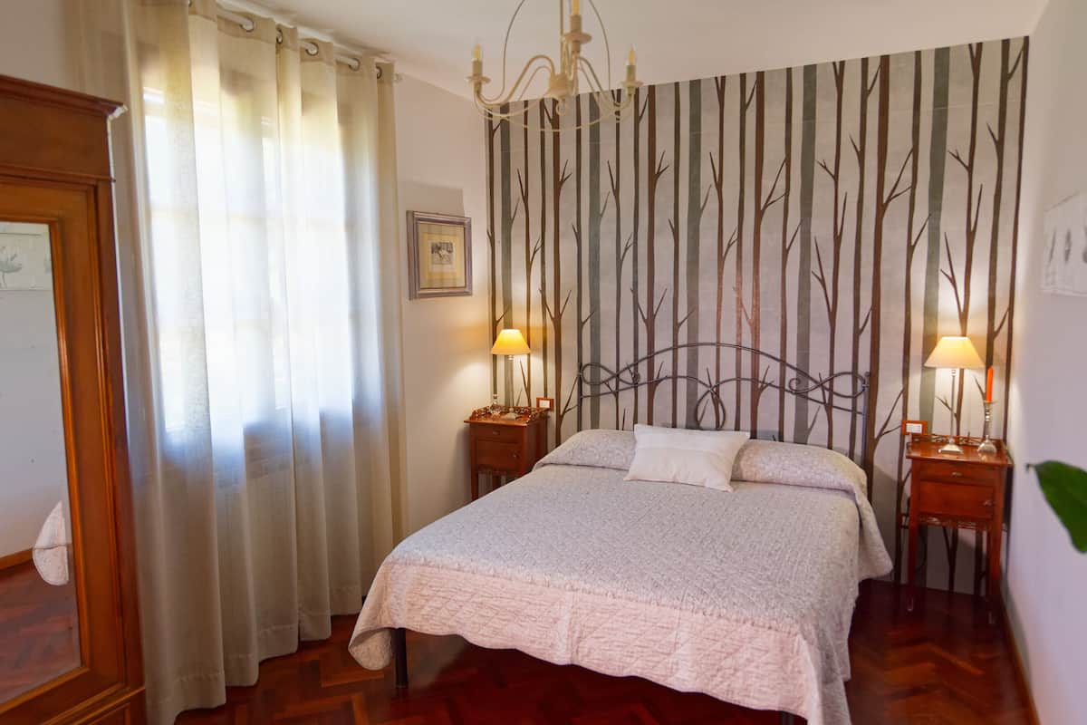 Une chambre à coucher avec un lit, deux tables de chevet avec des lampes, un lustre, une fenêtre avec des rideaux et un mur orné d’un motif d’arbre.
