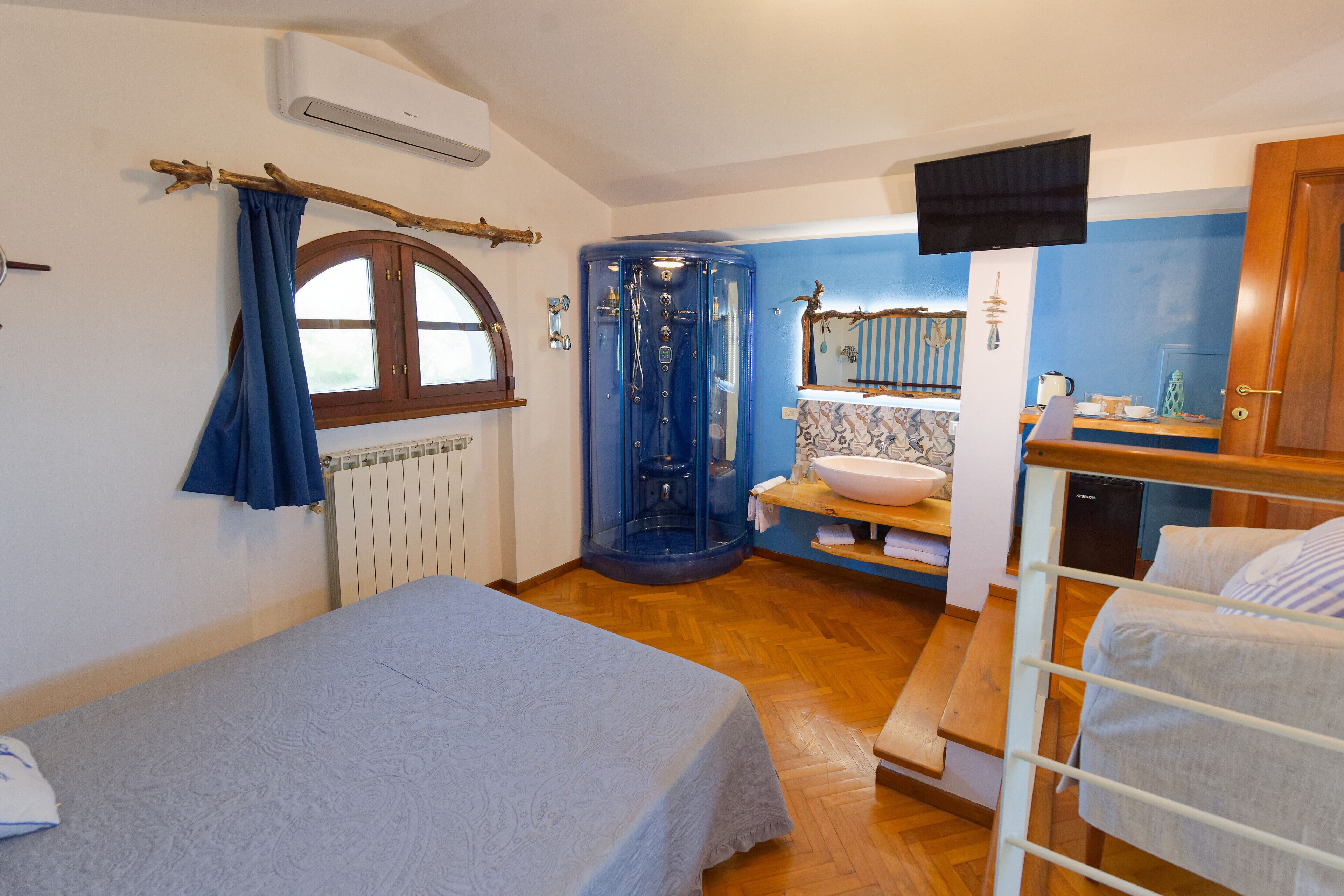 A bedroom with a bed, a wooden bench, a small table, a sink, and a shower enclosure.