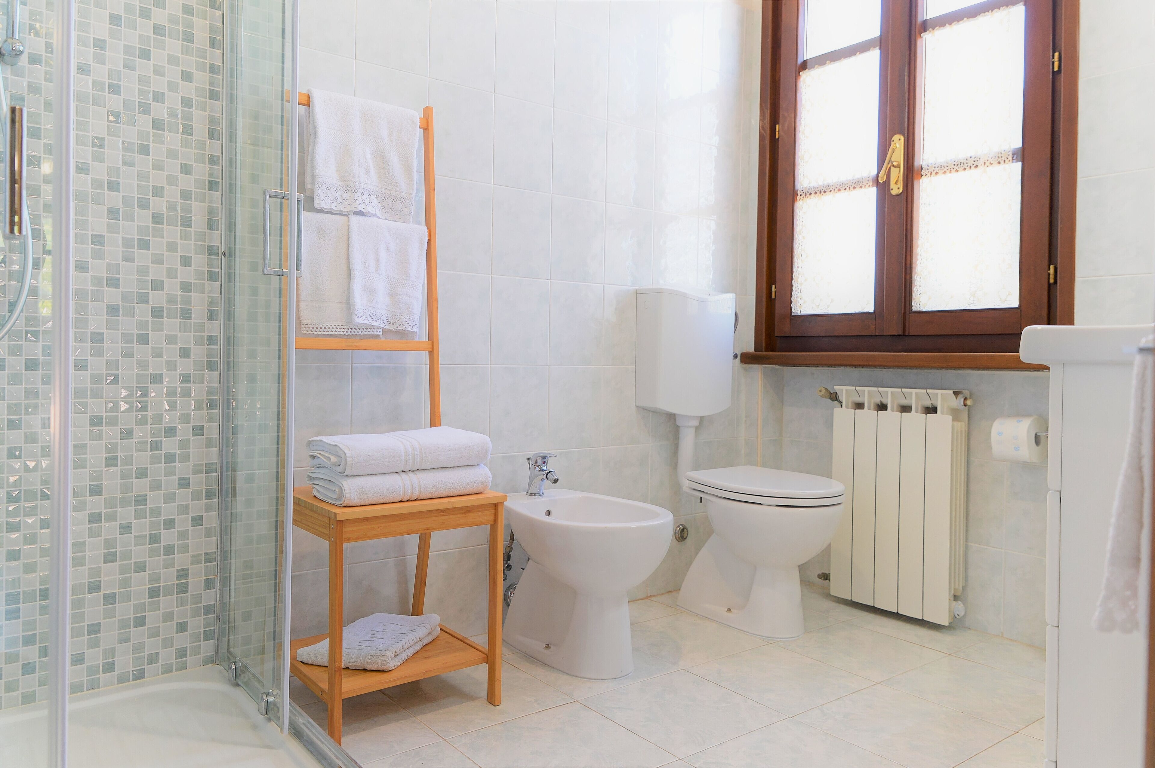 A bathroom with a glass shower enclosure, a wooden shelf with towels, a bidet, a toilet, and a radiator.