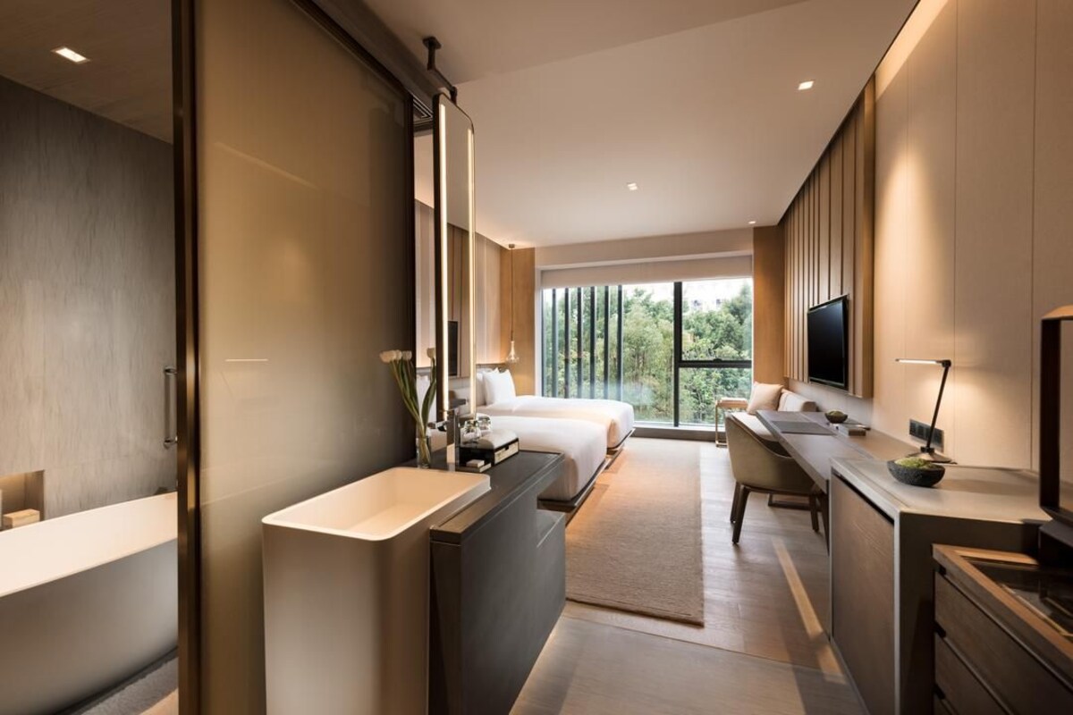 Quarto de hotel moderno com cama grande, área de estar separada, escrivaninha, televisão e janela ampla com vista para o exterior.