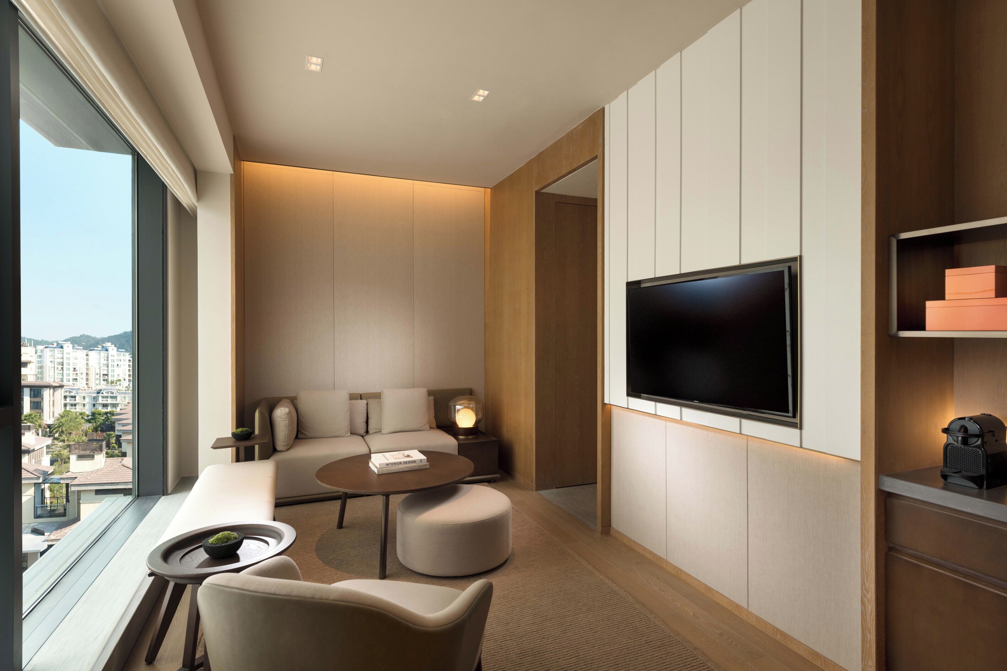 Habitación de hotel moderna con un televisor grande, asientos cómodos, una mesita y vistas a la ciudad.
