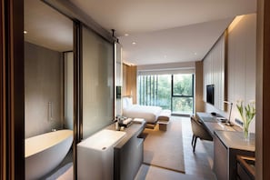 Quarto de hotel moderno com uma banheira grande, área de chuveiro separada, escrivaninha com cadeira, cama e uma janela ampla com vista para o verde.