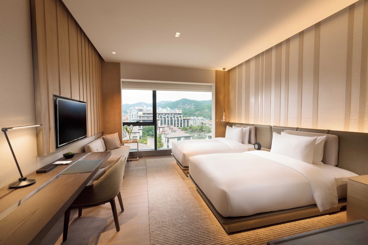 Quarto de hotel com uma cama grande, uma escrivaninha com computador, uma televisão e vista para a cidade.