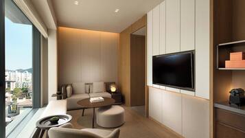 Quarto de hotel moderno com televisão grande, assentos confortáveis, uma mesinha e vista para a cidade.