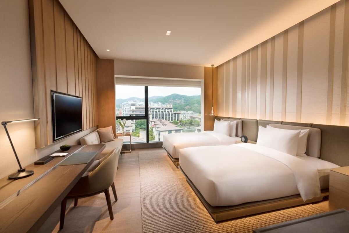 Quarto de hotel com uma cama grande, uma escrivaninha com cadeira, uma televisão e vista para a cidade através de amplas janelas.
