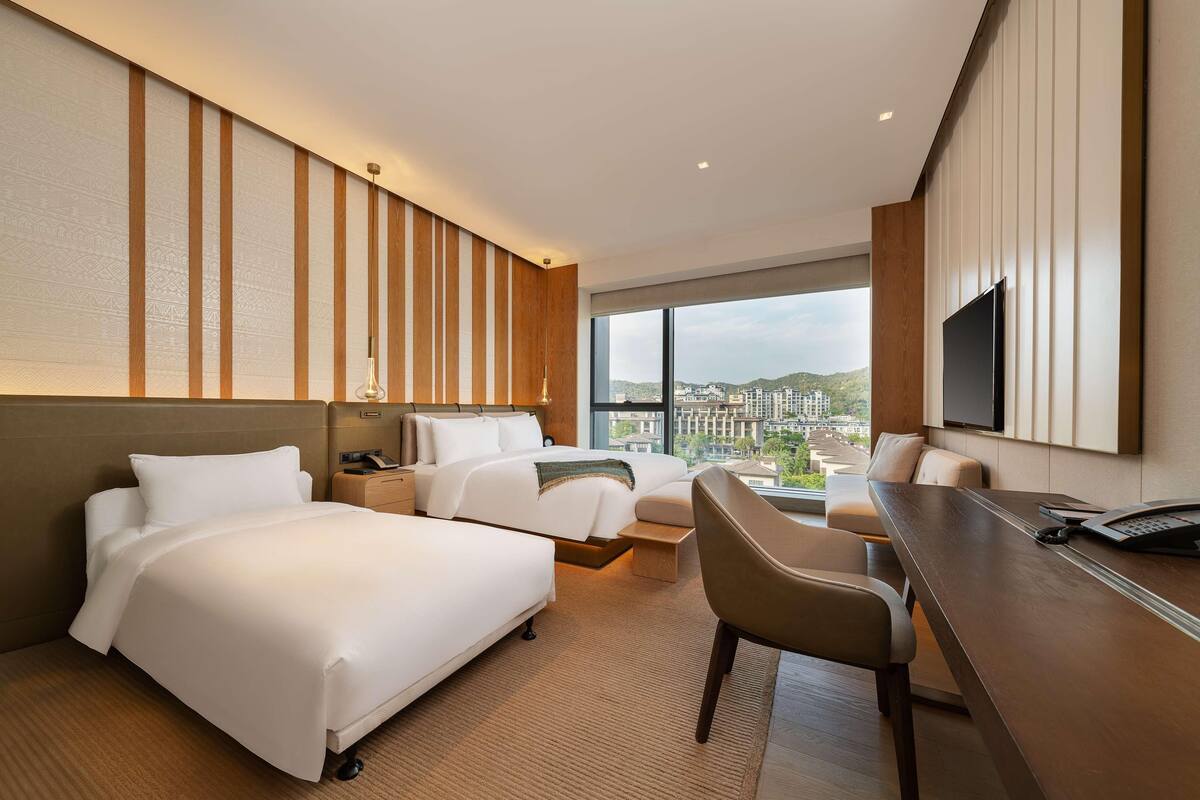 Une chambre d’hôtel avec deux lits, un bureau, une chaise, une télévision et une grande fenêtre donnant sur la ville.