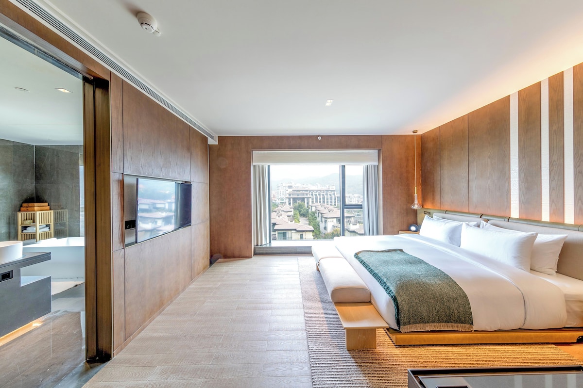 Quarto de hotel moderno com uma cama grande, televisão, vista para a cidade e porta deslizante para o banheiro.
