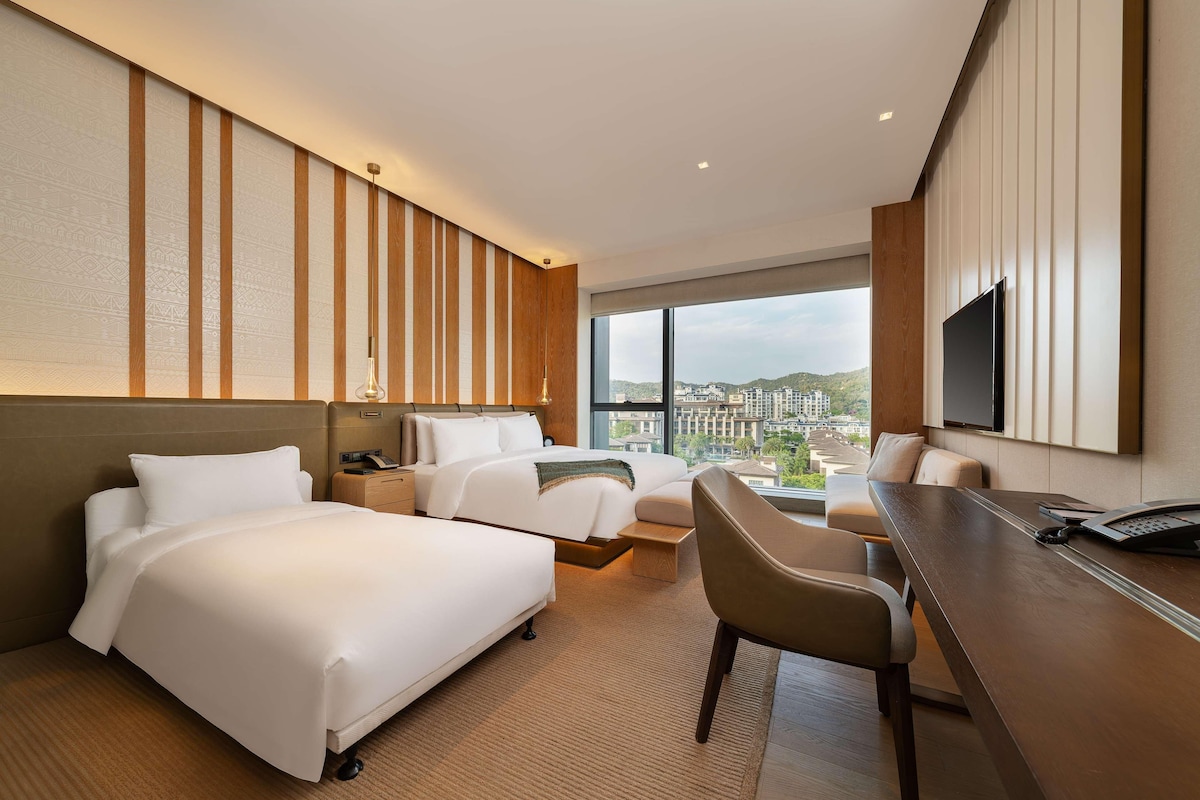 Quarto de hotel com duas camas, uma escrivaninha, uma cadeira, televisão e uma janela grande com vista para a cidade.