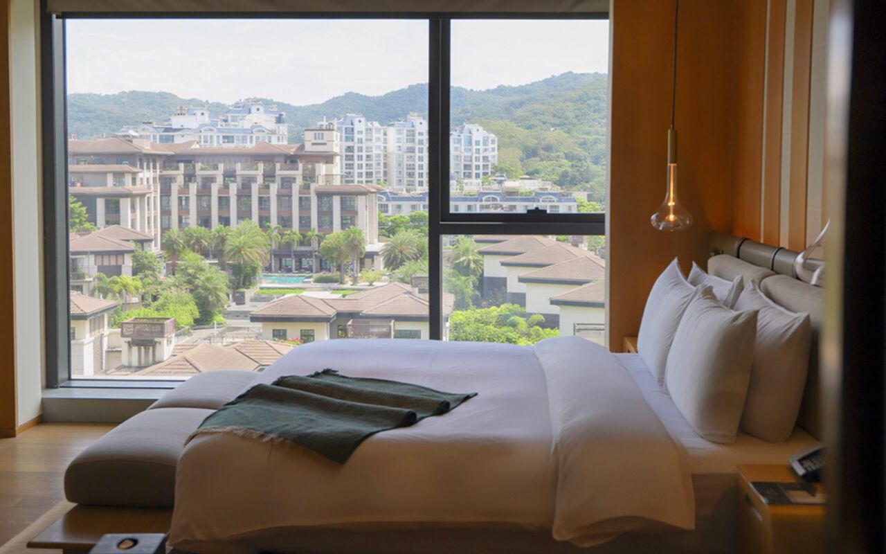 Habitación de hotel con una cama grande, vistas a edificios y vegetación, y una lámpara colgante.