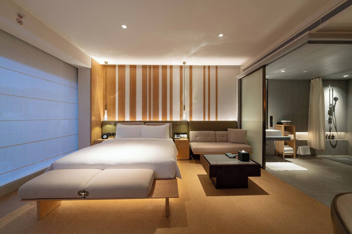 Une chambre d’hôtel moderne, dotée d’un grand lit, d’un canapé, d’une table basse, d’une table de chevet et d’une salle de bain visible à travers une paroi en verre.