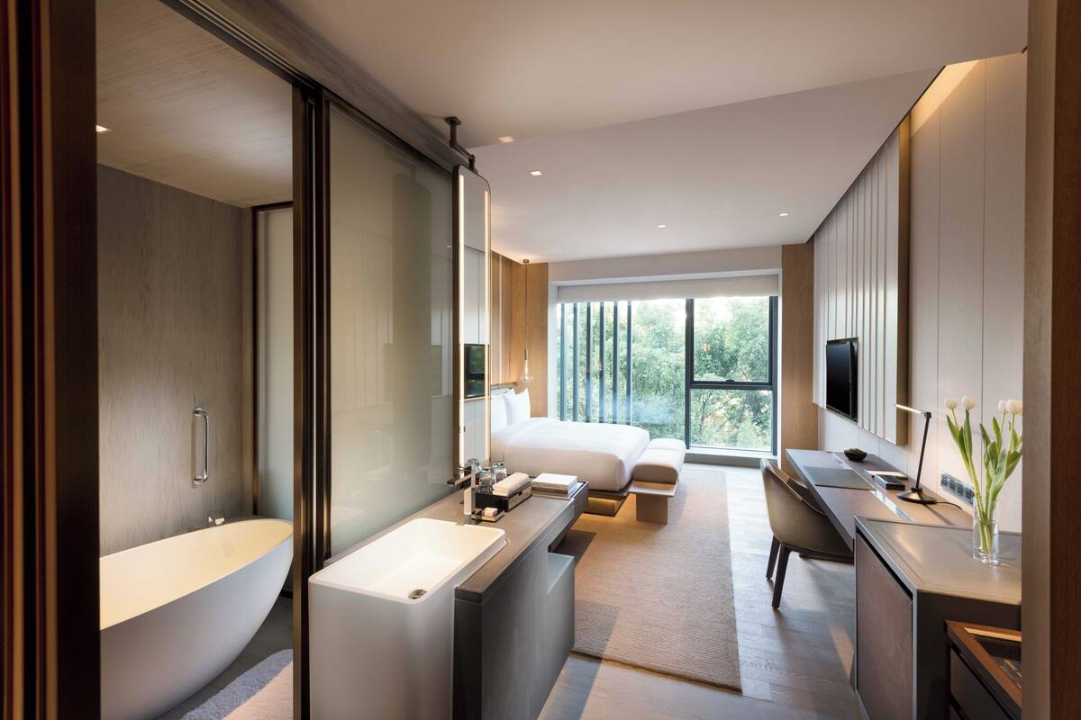 Une chambre d’hôtel moderne dotée d’une grande baignoire, d’une douche séparée, d’un bureau avec une chaise, d’un lit et d’une grande fenêtre donnant sur un paysage verdoyant.