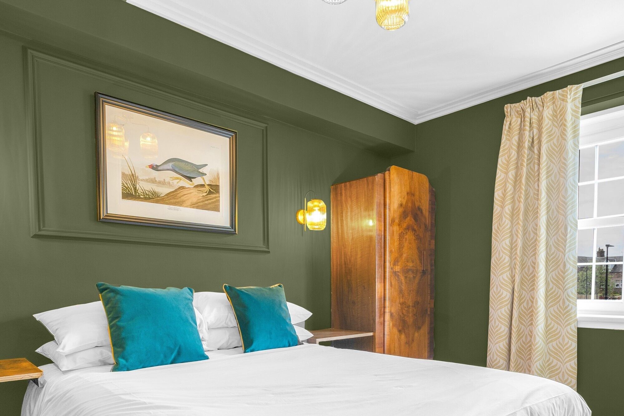 A bedroom with a bed, pillows, a wooden wardrobe, a framed picture, and a window with curtains.
