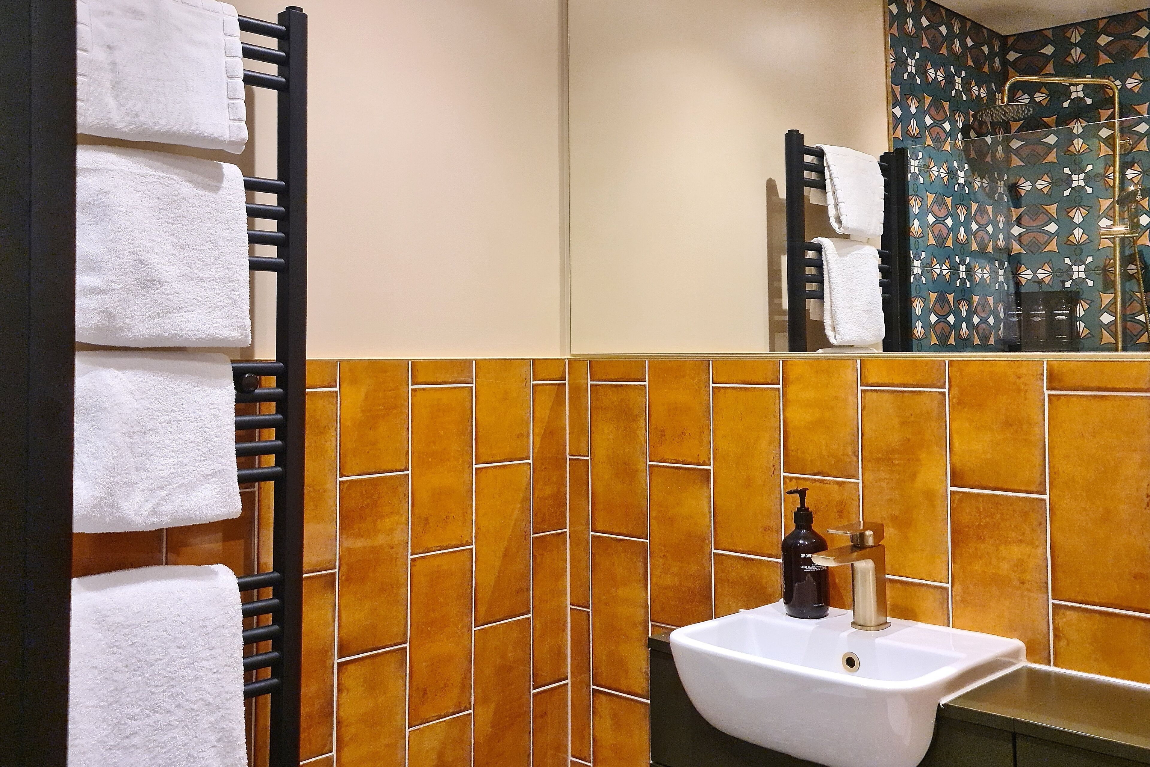 Un baño con pared de azulejos, un lavamanos con dispensador de jabón y un perchero con toallas blancas.
