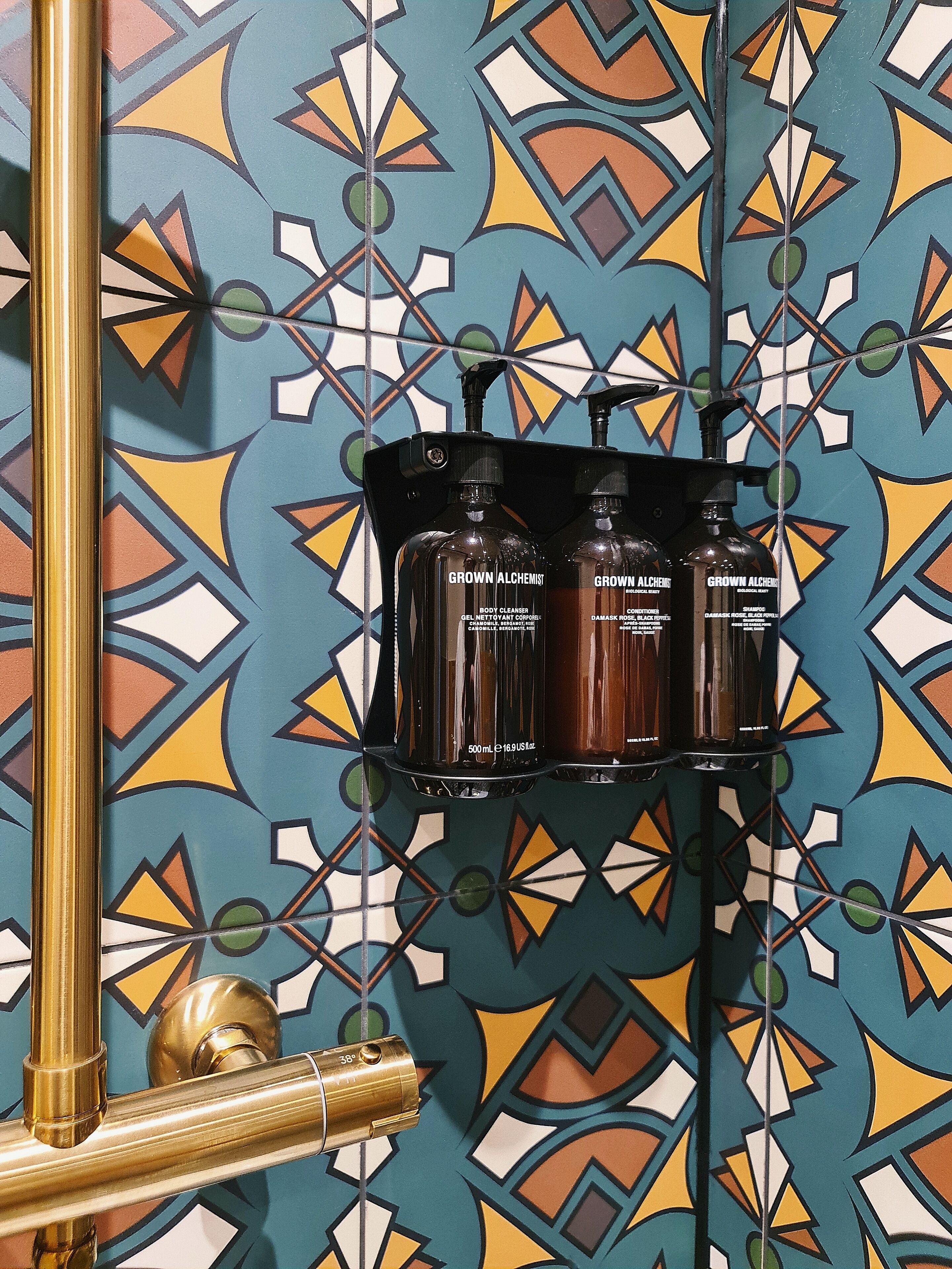 A bathroom with a geometric patterned tile wall, a gold-colored faucet, and a black shelf holding three bottles labeled 'GROWN ALCHEMIST'.