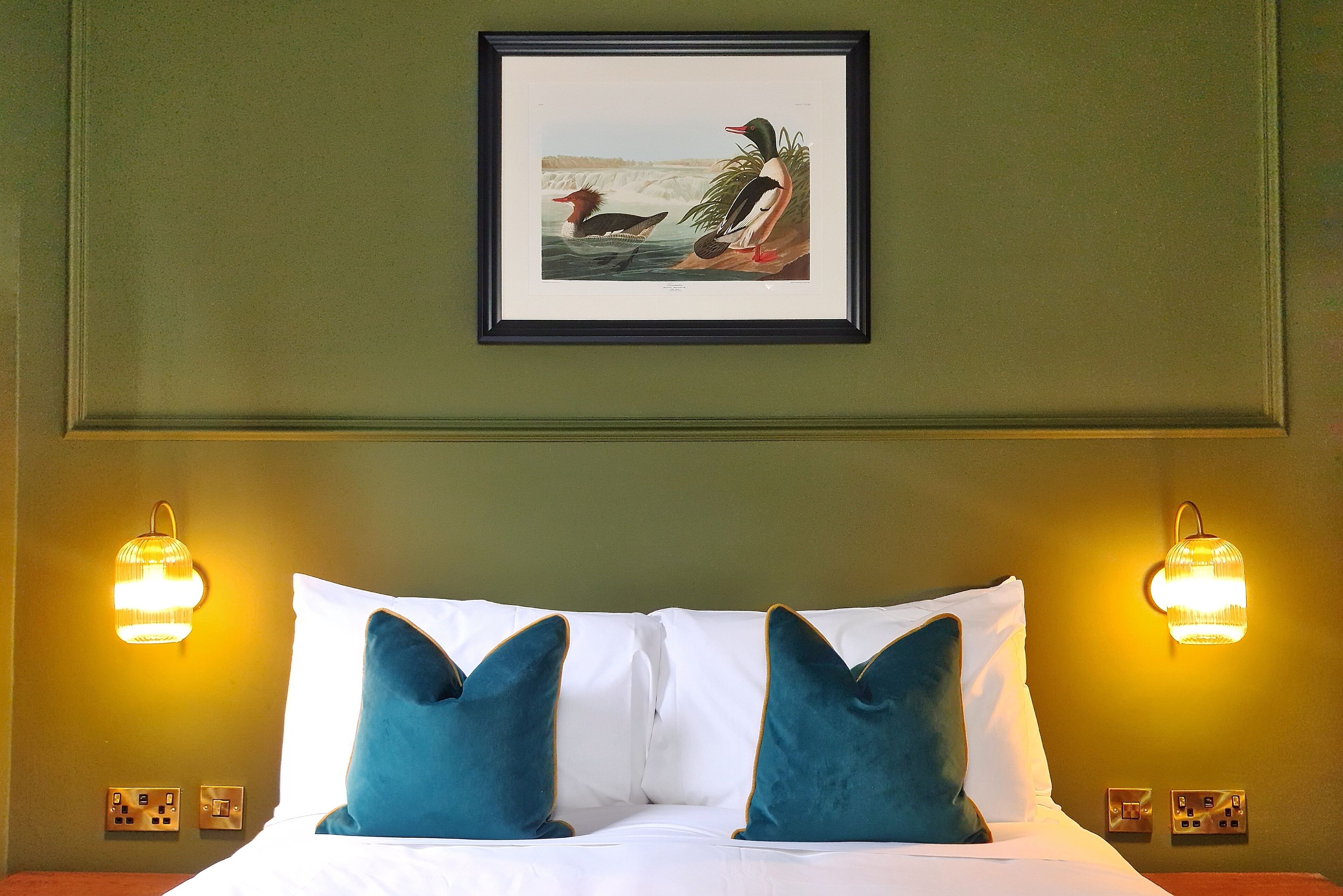 A hotel room with a bed, two teal pillows, a framed picture of ducks, and wall-mounted lights.