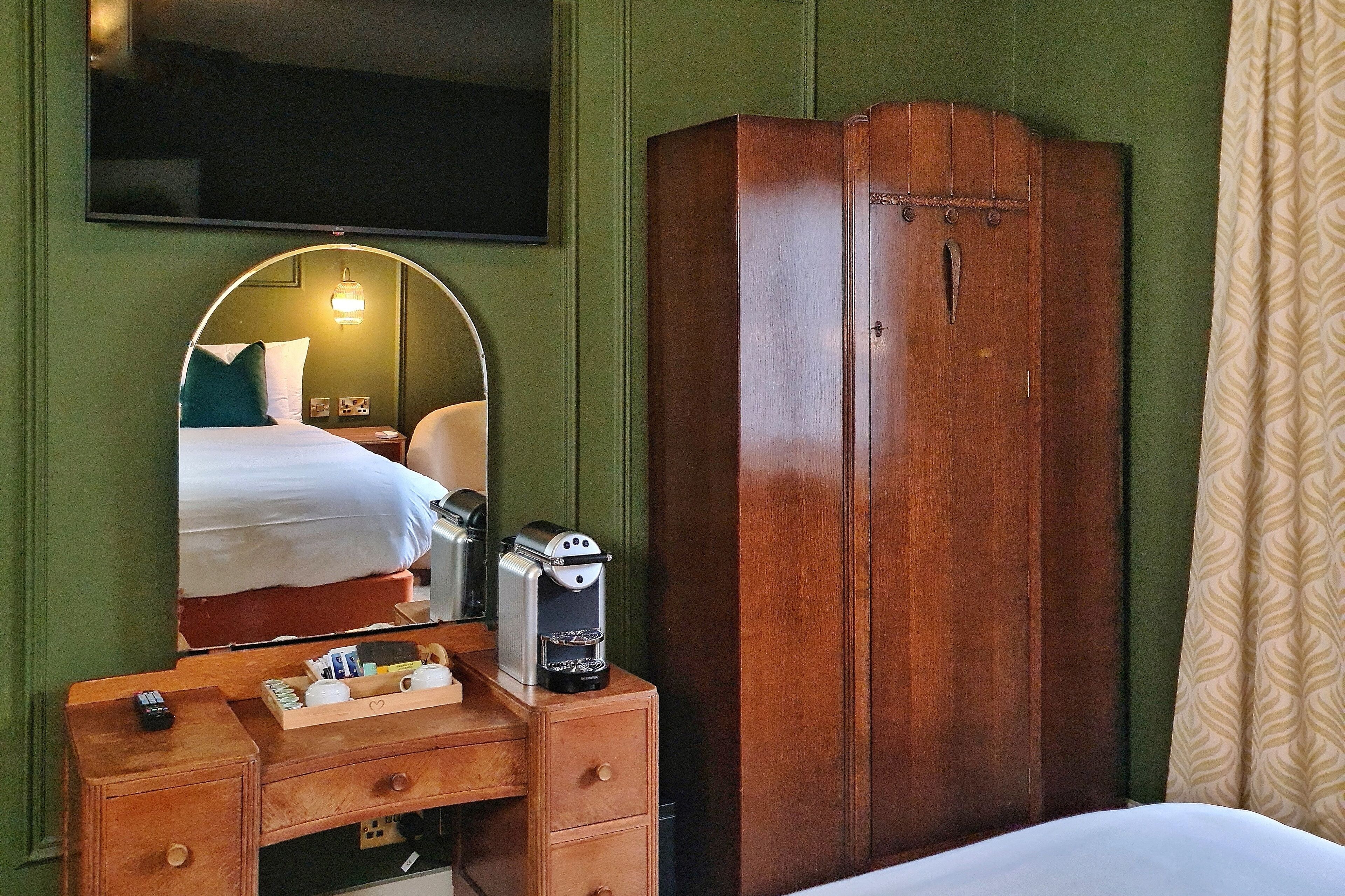 A hotel room with a bed, a wooden wardrobe, a bedside table with a coffee maker, and a mirror reflecting the bed and a wall-mounted TV.