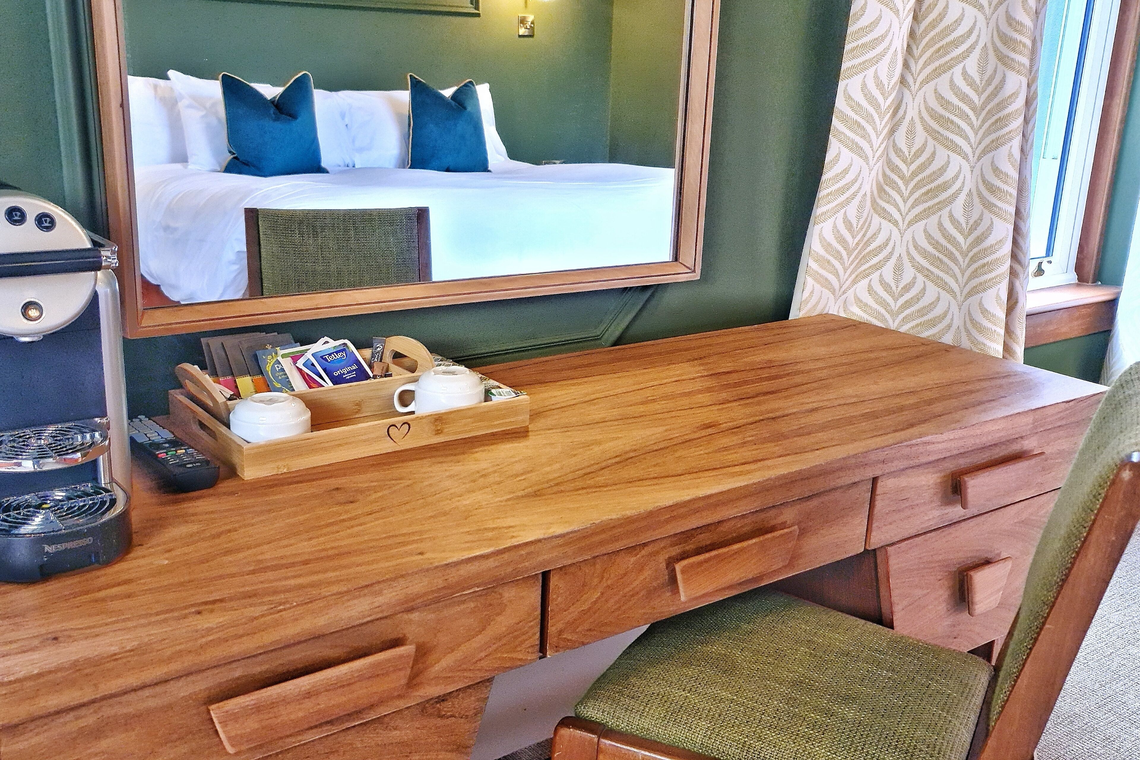 Una scrivania in legno con un vassoio che contiene una macchina da caffè, telecomandi e un libro. Uno specchio riflette un letto con cuscini blu.