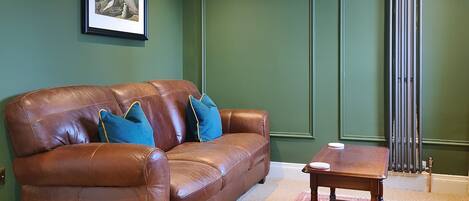 A brown leather sofa with blue cushions, a wooden side table, a framed picture on the wall, and a radiator.