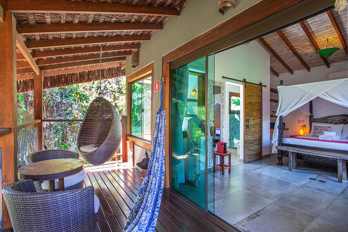 Una terraza de madera con muebles de mimbre y una hamaca, que conduce a una puerta de cristal hacia una habitación con cama y mesita de noche.