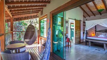 Una terraza de madera con muebles de mimbre y una hamaca, que conduce a una puerta de cristal hacia una habitación con cama y mesita de noche.