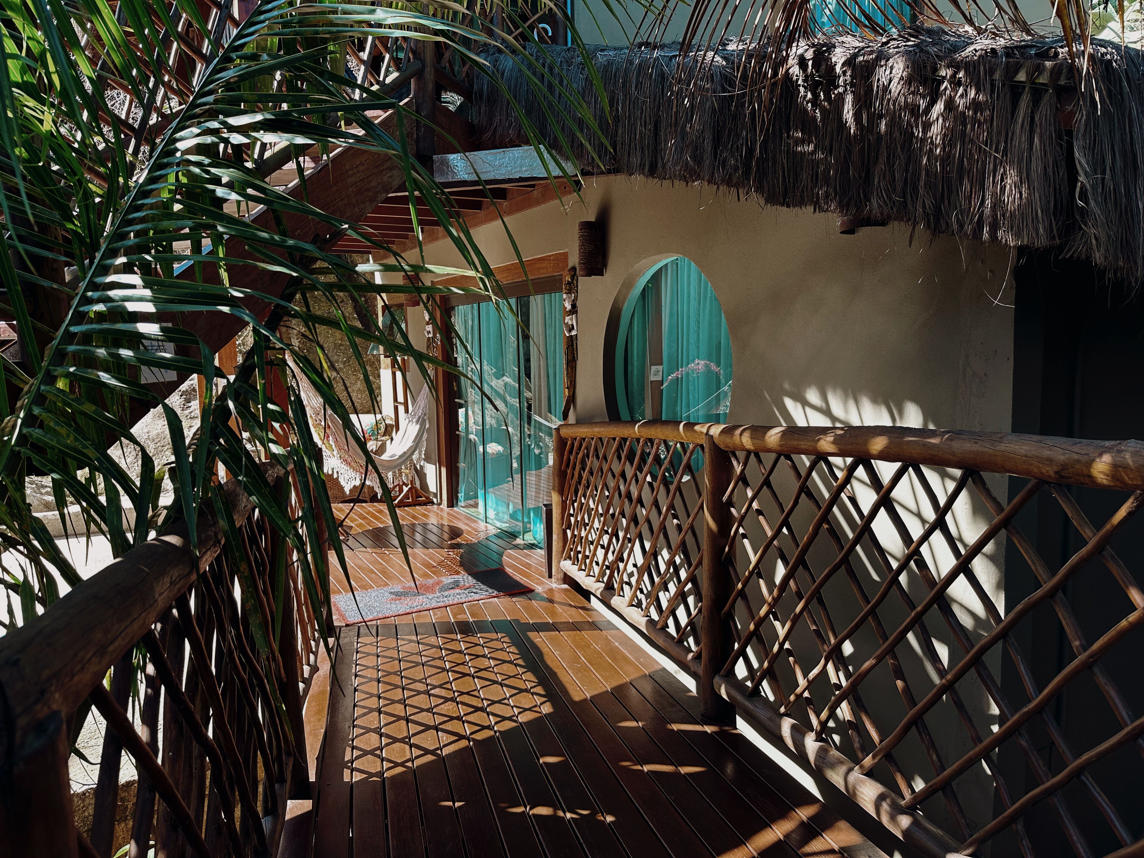 Une terrasse en bois surmontée d’un toit de chaume, qui mène à un bâtiment doté d’une porte verte.