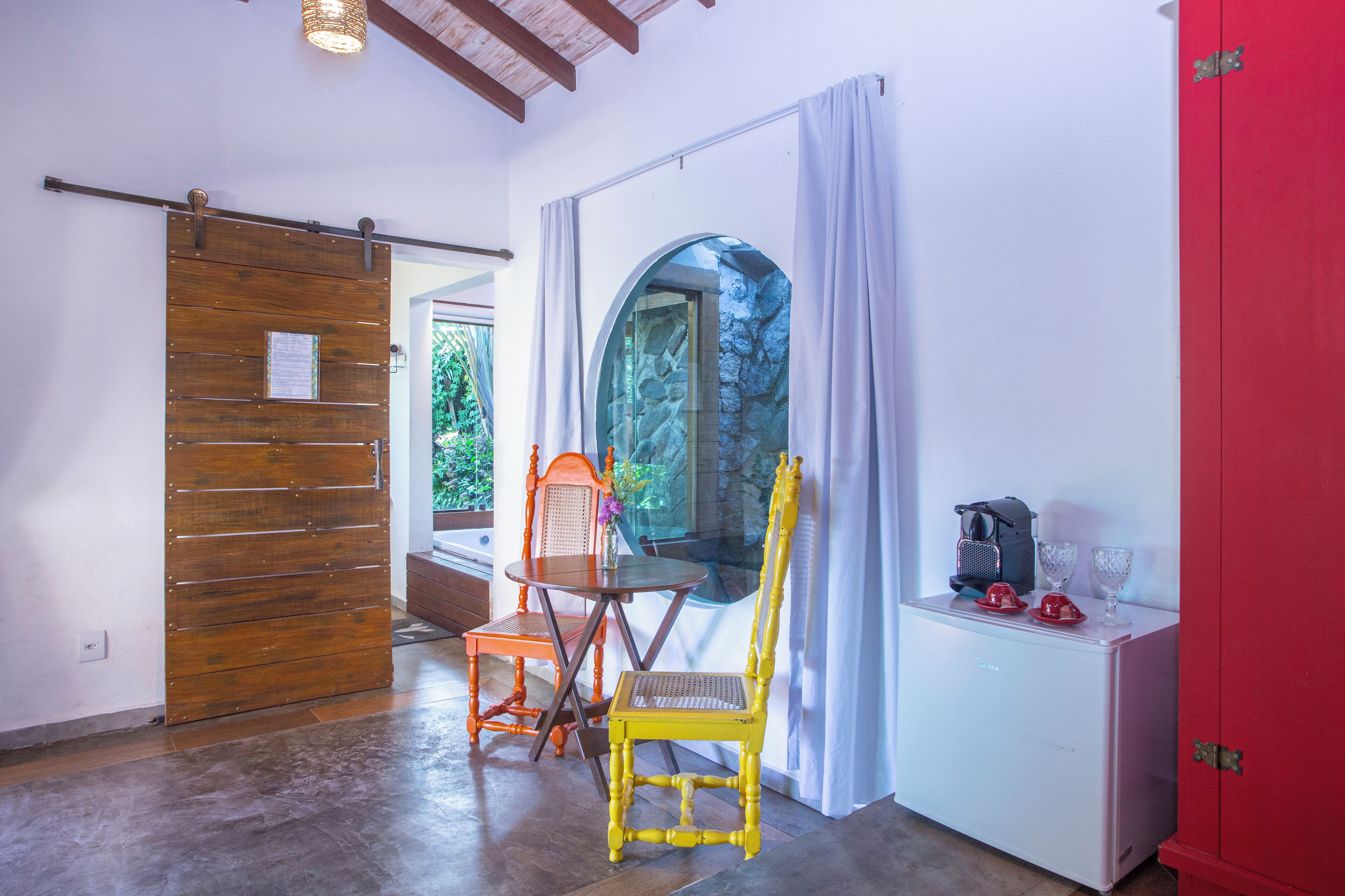 Una habitación con puerta corredera de madera, ventana con vistas a follaje, una silla amarilla, un armario rojo y una mesita con cafetera y vasos.