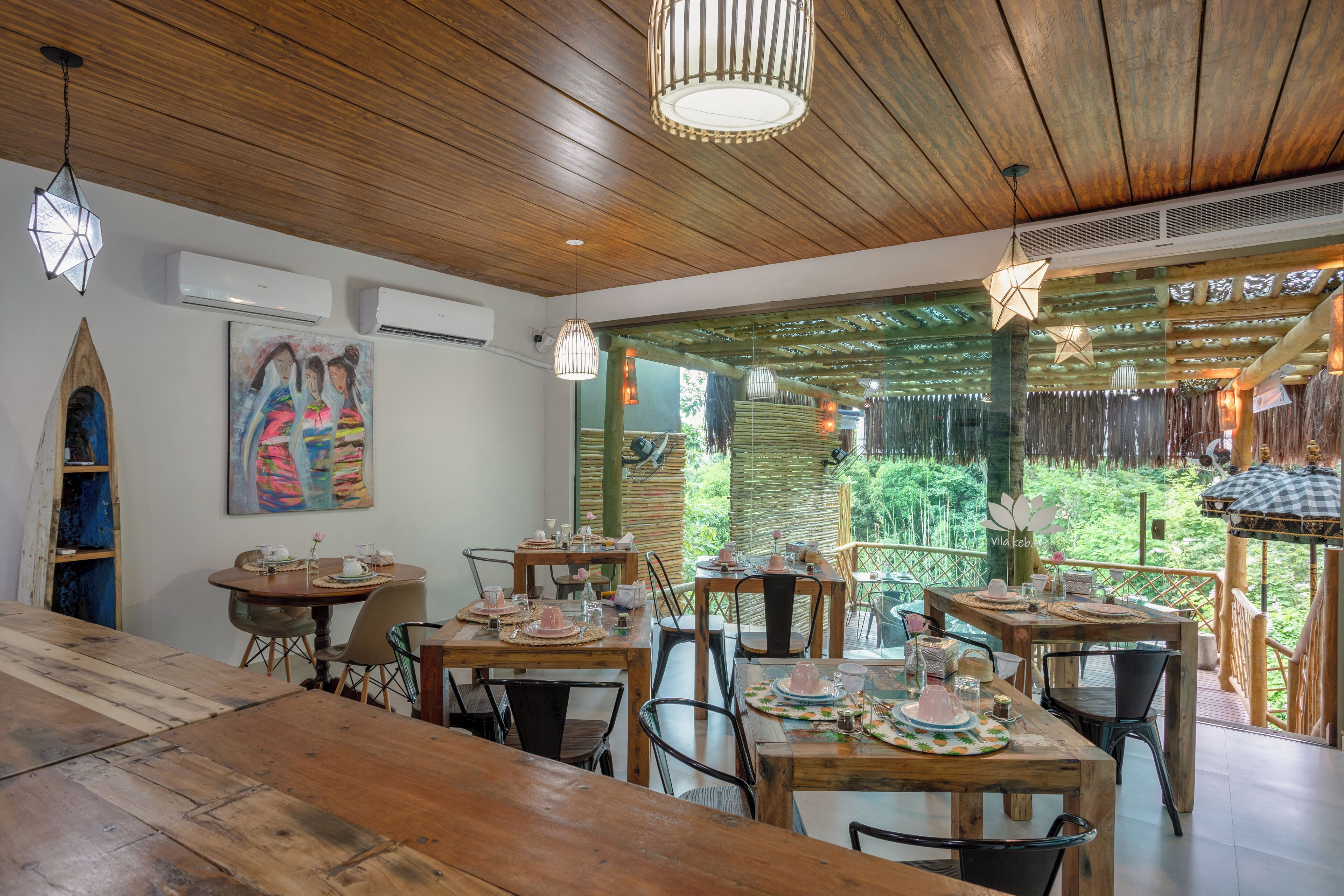 Un comedor con mesas y sillas de madera, un cuadro grande en la pared y vistas al exterior con vegetación.