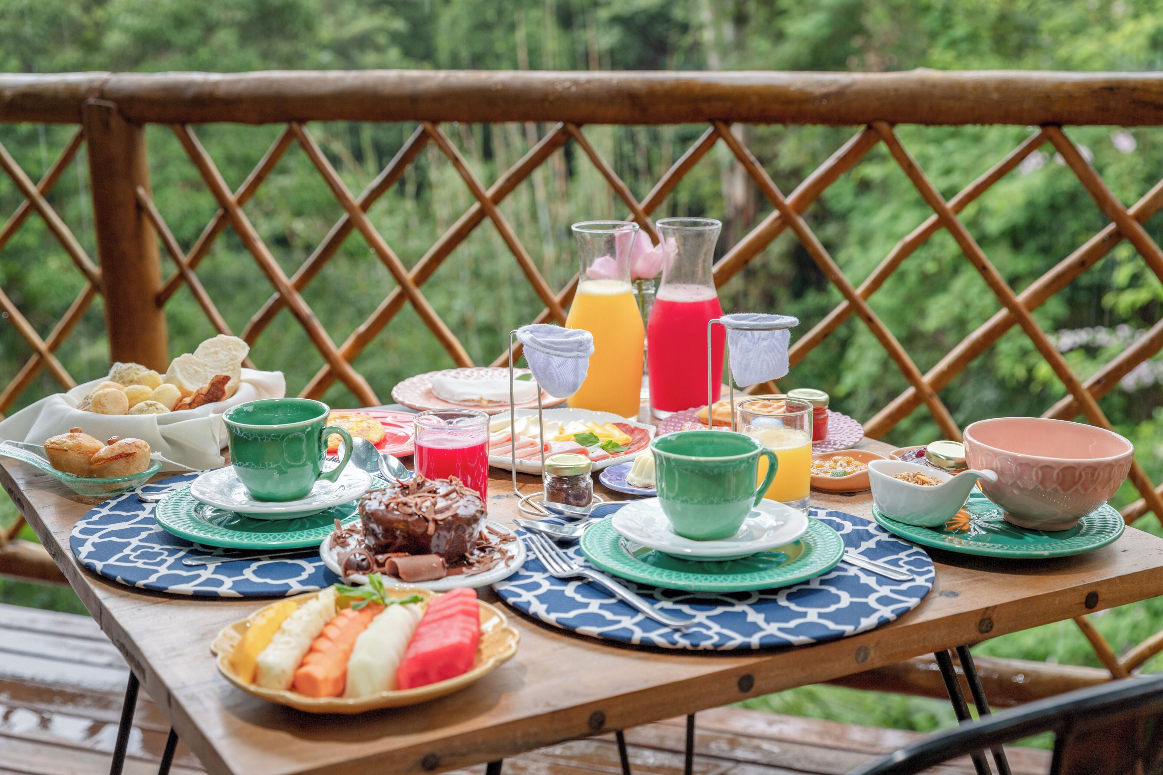 Ett träbord dukat med frukosttillbehör, inklusive bakverk, frukt och drycker, på en balkong med ett bamburäcke och frodig grönska i bakgrunden.
