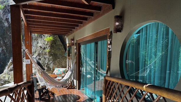 A wooden deck with a hammock, a wooden table, and a view of a rocky landscape through a glass door. - Hotel Vila Kebaya (Ilhabela)