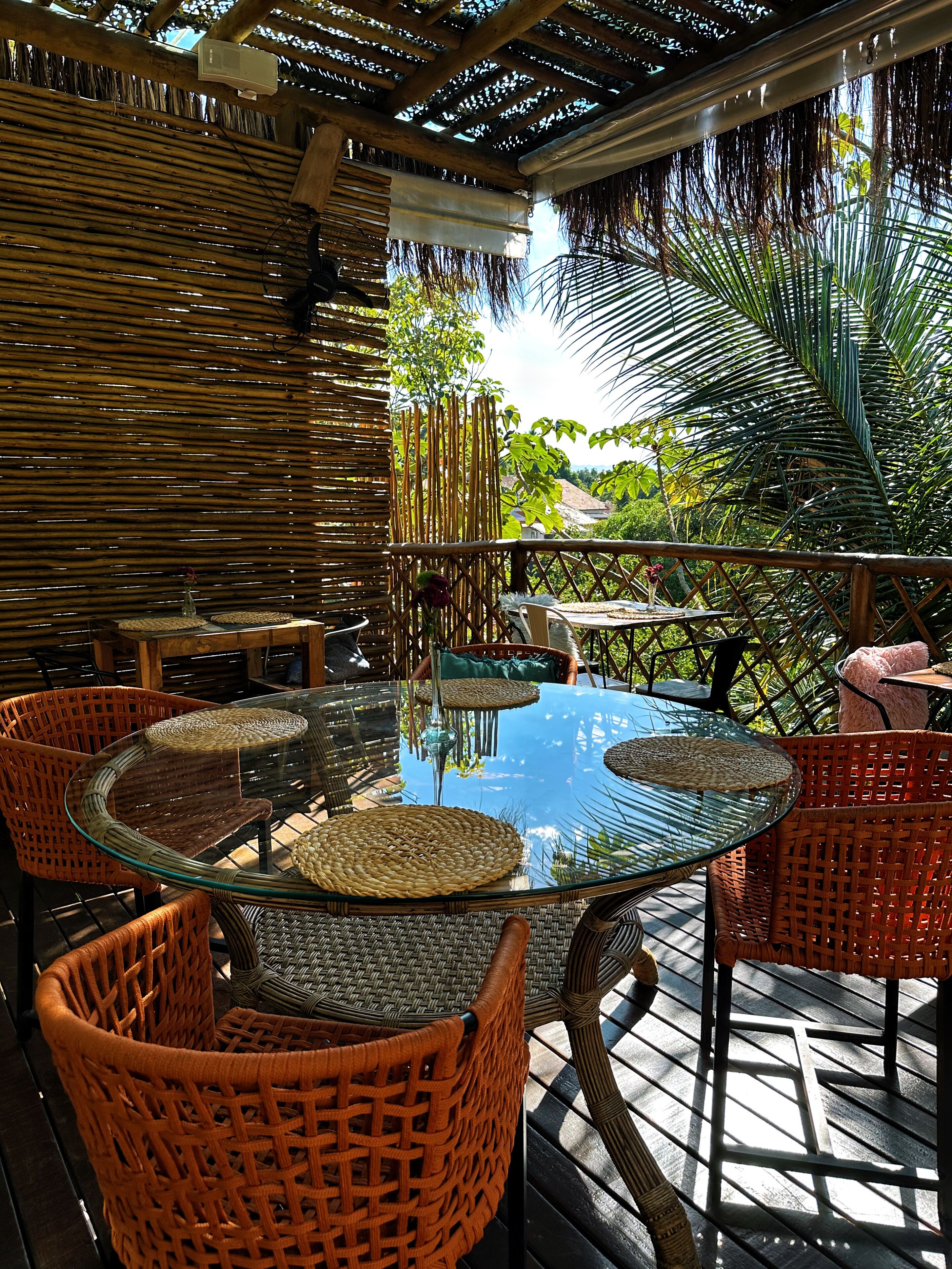 An outdoor patio with round glass tables and wicker chairs, surrounded by a bamboo fence and tropical plants.