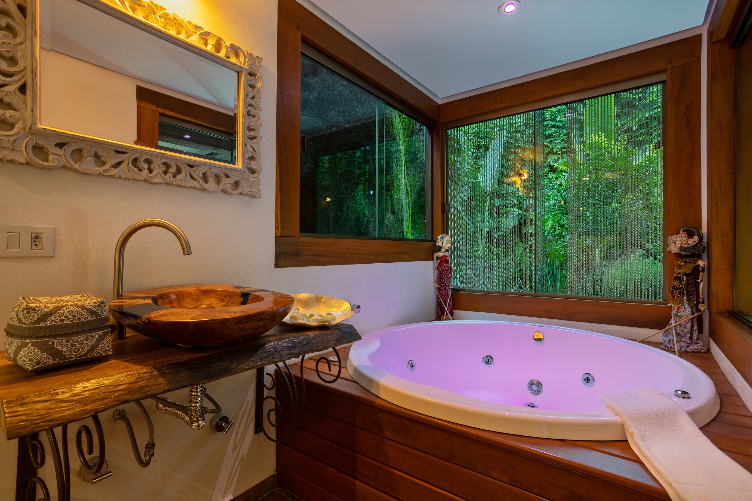 Un baño con tocador de madera, una bañera ovalada grande, un espejo con marco ornamentado y una ventana con vista a follaje.