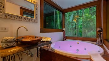 Un baño con tocador de madera, una bañera ovalada grande, un espejo con marco ornamentado y una ventana con vista a follaje.
